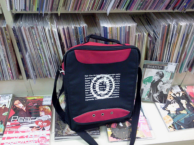 Color photo of OTW tote bag resting on a counter directly in front of a display of doujinshi on shelves.