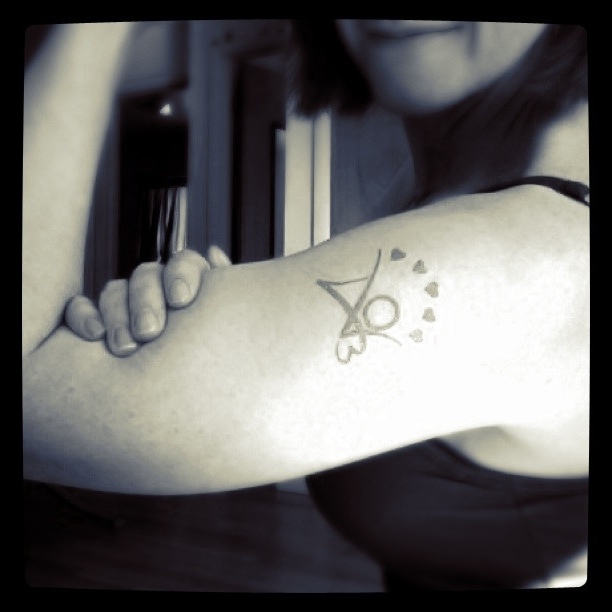 Black and white photo showing woman with face out of frame, raising arm with AO3 kudos temporary tattoo displayed on bicep.