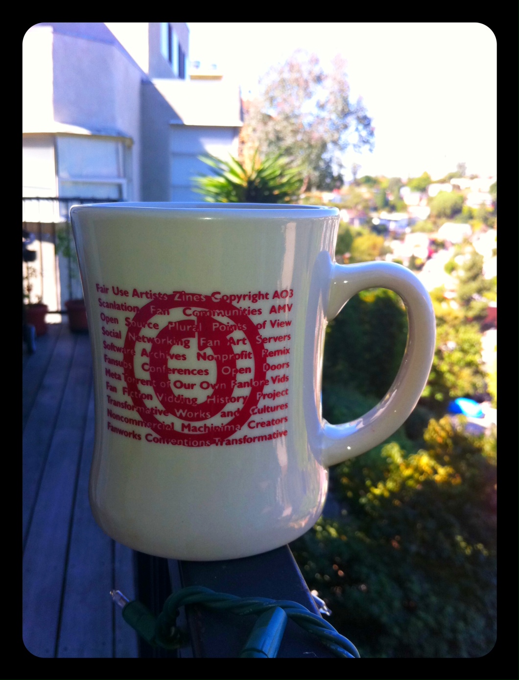 Color photo of OTW ceramic diner mug sitting in shadow on balcony railing, with sunlit trees in the background.