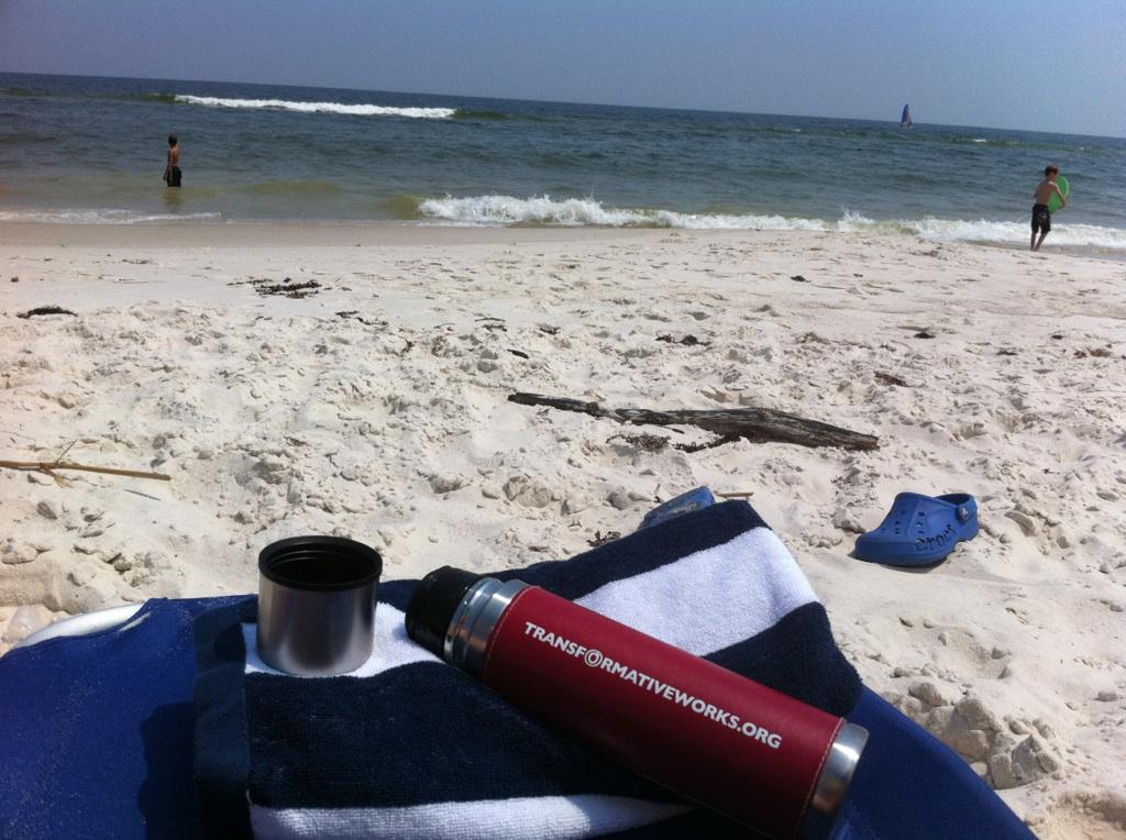 Color photo of sandy beach with OTW thermos resting on a beach towel. A lake or sea is in the background, with waves cresting.