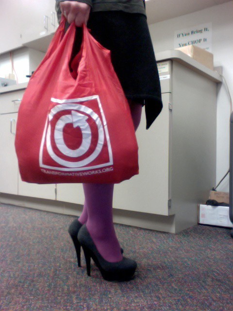 Color photo of OTW nylon carryall carried by a person wearing high heels, showing only their hand and legs. There are office cabinets in the background.